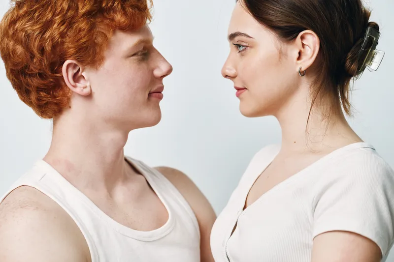 Young man and woman facing each other with neutral expressions, closeup portrait, casual clothing, studio light background, intimate moment and connection.