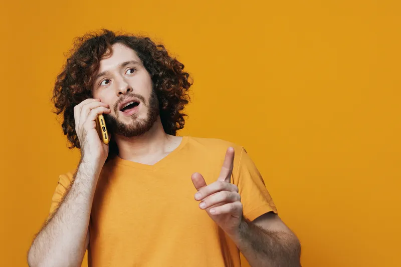Young man with curly hair talking on the phone against a bright yellow background, expressing surprise and excitement, embodying modern communication