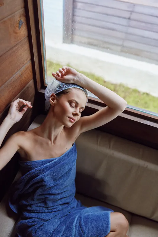 Relaxed woman wrapped in blue towel and wearing shower cap sits near window in wooden sauna, enjoying calm moment with closed eyes and soft natural light around.