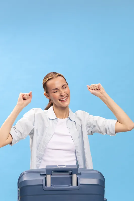 Happy young woman with suitcase raises arms in celebration, smiling with closed eyes. Blue background. Concept of travel, vacation, excitement, and freedom for tourists and travelers.