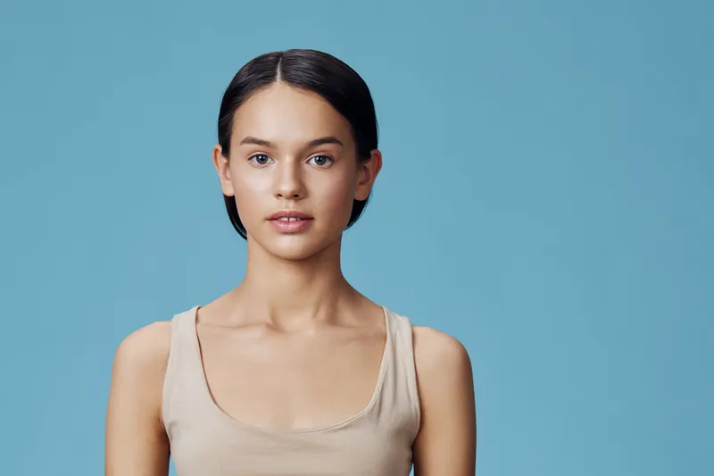 Young woman with clear skin and a neutral expression against a blue background, showcasing natural beauty and simplicity in a minimalist style