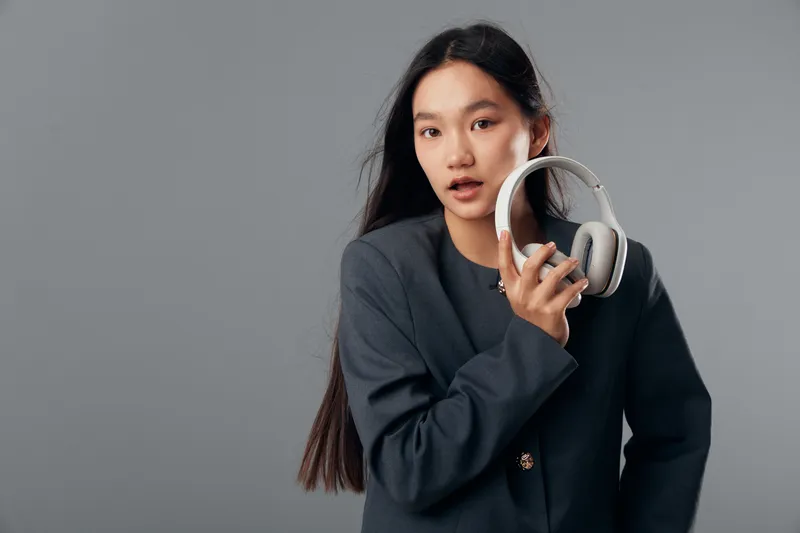 Young woman with long black hair in dark jacket holding white headphones on gray background for music, technology, communication, lifestyle and entertainment concept portrait.