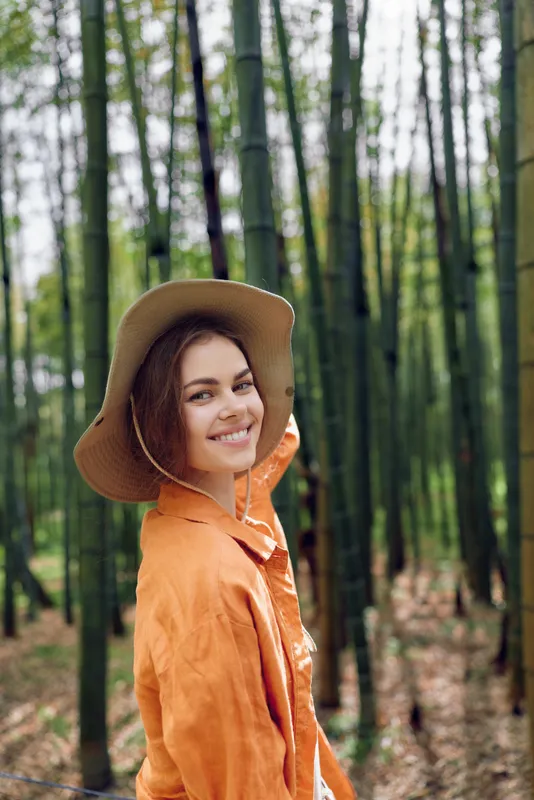 Woman portrait outdoors in a bamboo forest for travel, smiling woman wearing a hat and orange jacket, natural sunlight and happy smile among tall green trees.