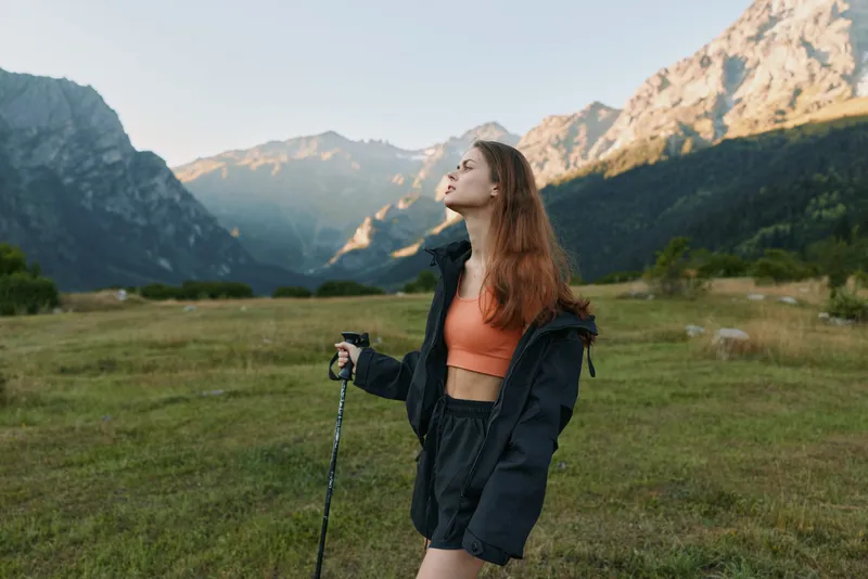 A young woman stands in a grassy valley beneath towering mountains, dressed for hiking in a windproof jacket and orange top, gazing upward with a sense of exploration.