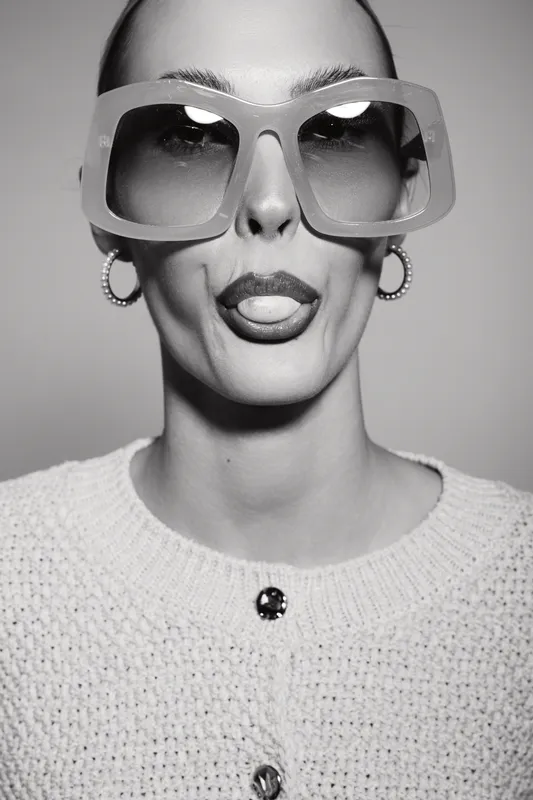 Portrait of a caucasian woman with pale skin wearing large sunglasses and a white sweater sticking her tongue out. Studio black and white photo with playful expression.