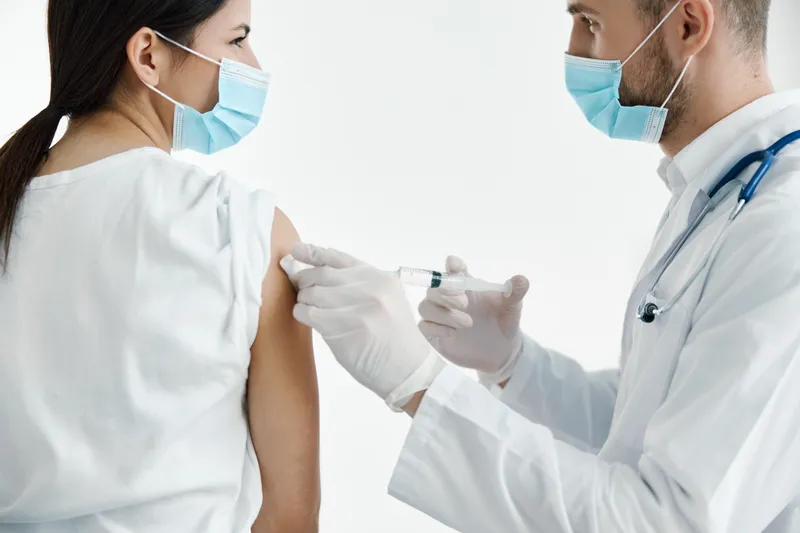 Врач в белом халате делает женщине укол в плечо вакциной, используя защитные перчатки
