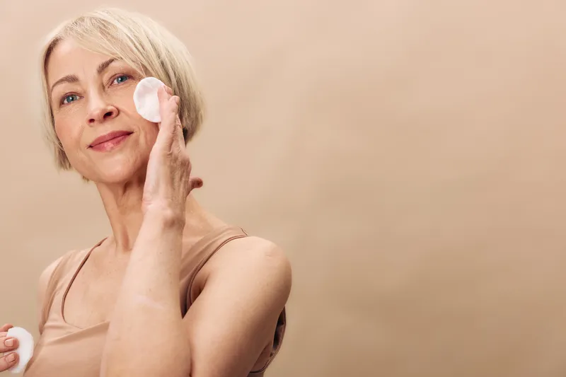 Mature woman with short blonde hair applies facial cream with cotton pad in bright studio. Concept of skincare, beauty routine, self-care, and healthy aging with soft natural lighting.
