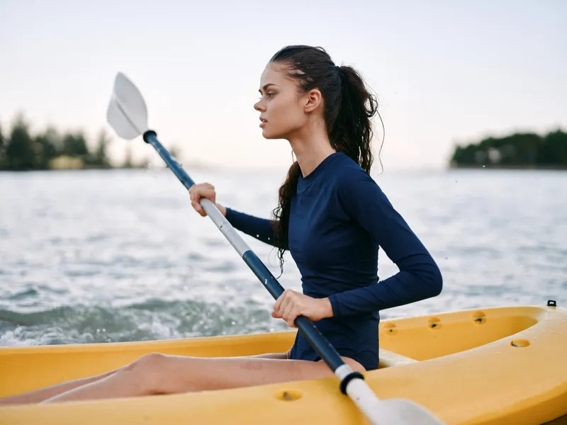 Athletic woman paddling a yellow kayak on calm water, wearing a dark long sleeve top, focused expression as she uses a double blade paddle.