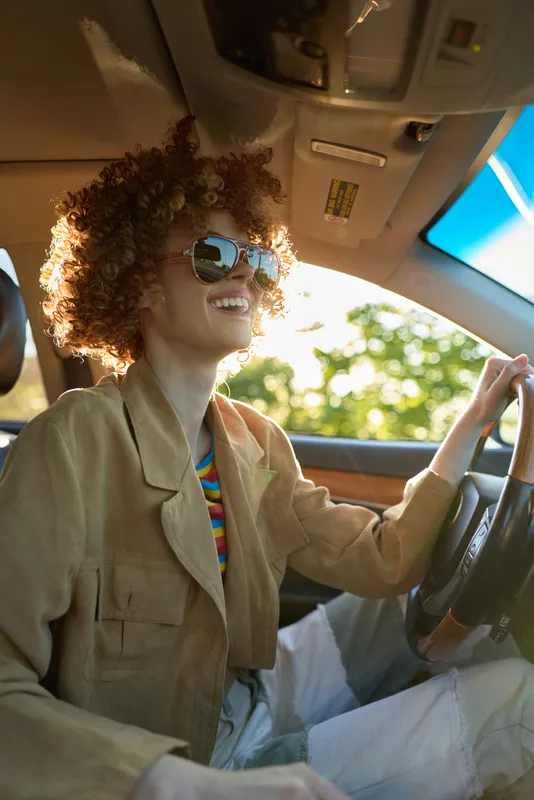 Woman driver with curly hair wearing sunglasses and a beige jacket sits at the wheel, smiling as sunlight fills the retro car interior, casual daytime lifestyle scene
