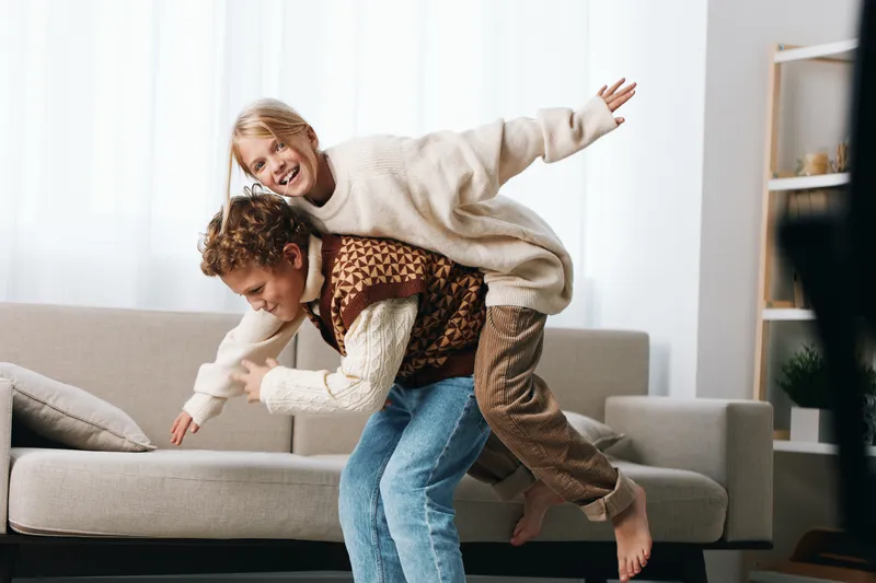 Playful siblings engage in a joyful indoor piggyback ride on a couch, in a bright living room, capturing carefree energy, laughter, and warm family moments.
