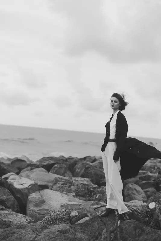 Woman stands on a rocky coast with a windblown coat, blackandwhite fashion photography on the sea horizon, moody outdoor landscape and ocean breeze with dramatic lighting and movement
