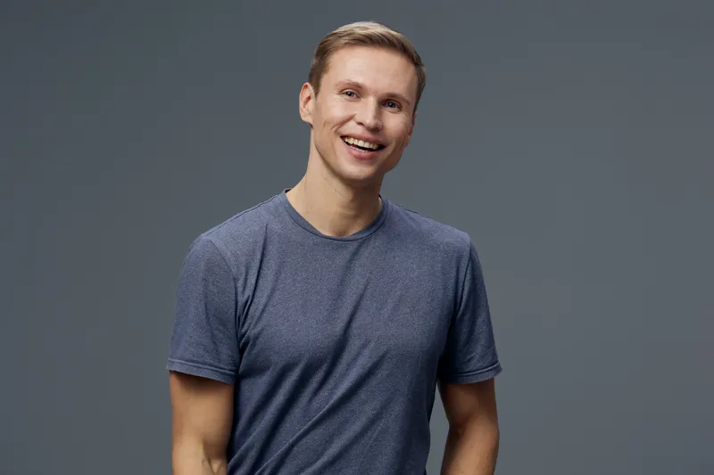 Smiling young man with short light brown hair wearing casual blue t-shirt looking at camera isolated on plain gray background studio portrait. People lifestyle concept, positive mood, leisure, relaxat