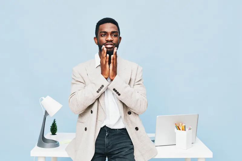Confident young man standing at desk touching his beard with thoughtful expression, casual beige blazer, white shirt, dark jeans, modern workspace with laptop, lamp, and plant, indoor studio shot, lif