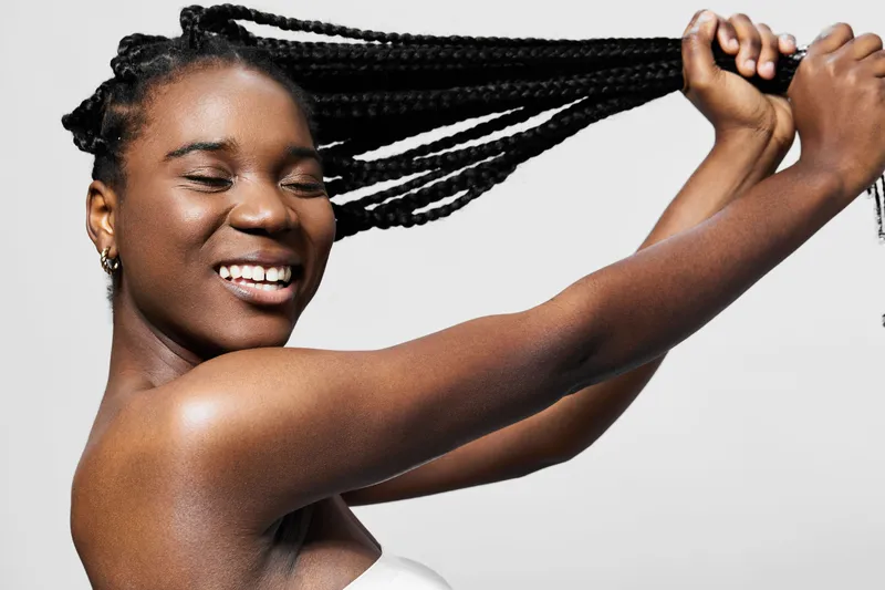 Happy Black woman with braided hair smiling while holding her hair, lifestyle focused on self expression and confidence, bright and clean background