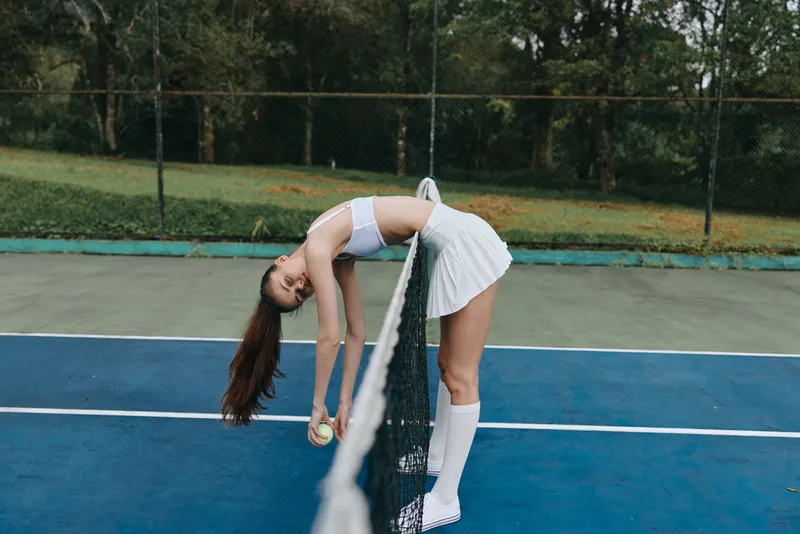 Young athletic female tennis player in sporty outfit stretching over the net, showcasing agility and determination on a sunny day in a vibrant tennis court