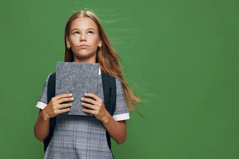 Girl with long hair holding closed book and wearing backpack standing on green background looking up. Child in casual clothes with thoughtful expression in studio with copy space.
