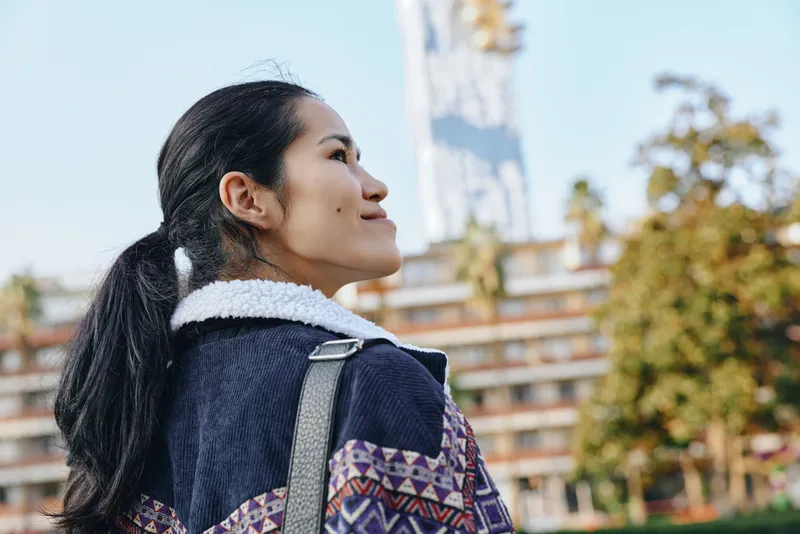 Woman looking outdoors in an urban setting with a patterned jacket and backpack, capturing casual fashion, travel mood, sunny daylight and street style during daytime exploration