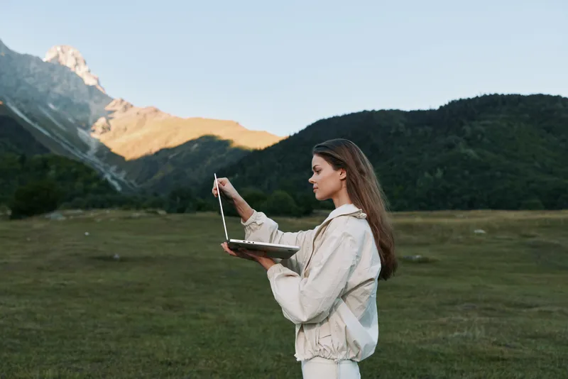 A focused woman stands outdoors with a laptop in a green meadow beneath distant mountains. She concentrates on her work, blending technology with nature to convey a calm, productive moment.