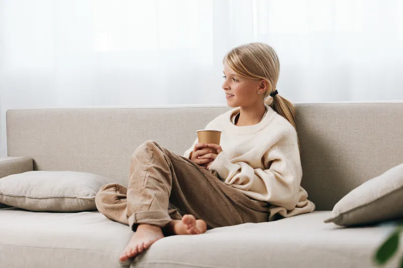 Authentic lifestyle image of a young girl resting on a sofa with a warm cup, wearing casual home attire, conveying a school themed moment at ease.