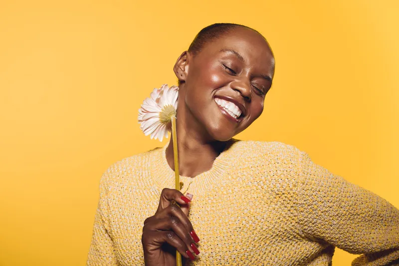 Smiling African woman with dark skin holding a flower near her face wearing a yellow sweater on a bright yellow background. Concept of happiness, beauty, and natural skincare.