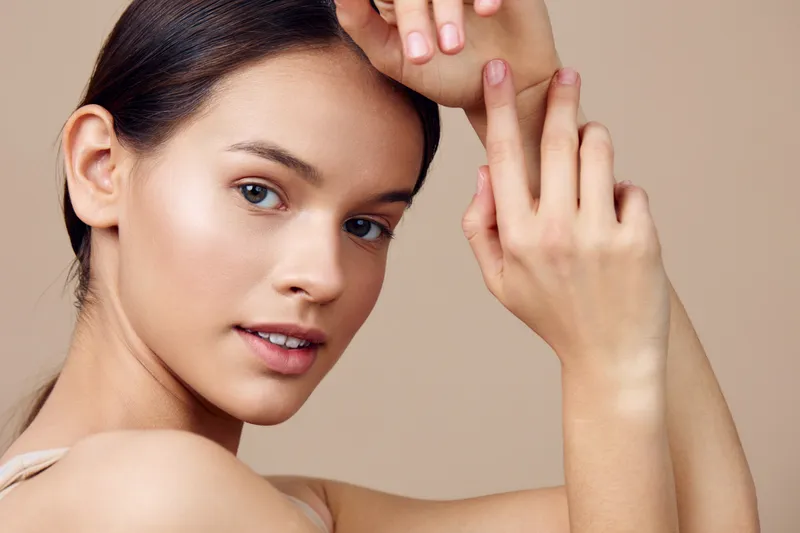 Portrait of a beautiful young model with natural makeup, showcasing glowing skin and delicate hands against a soft beige background
