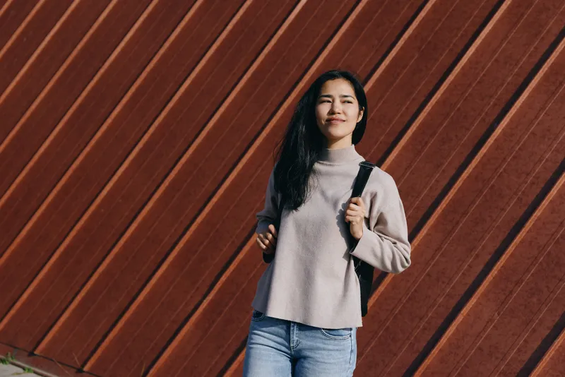 Woman with backpack by a diagonal wooden wall, wearing a cozy sweater and casual attire, urban street vibe, confident stance, daytime light, candid lifestyle shot for fashion, travel, and streetwear