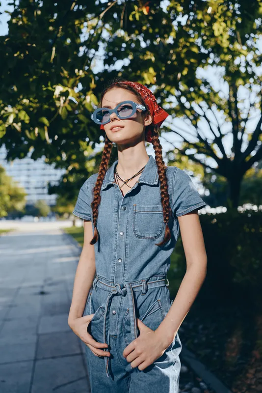 Modern boho-western denim street style woman wearing sunglasses and red bandana, standing confidently outdoors in bright film color lighting and casual summer outfit.