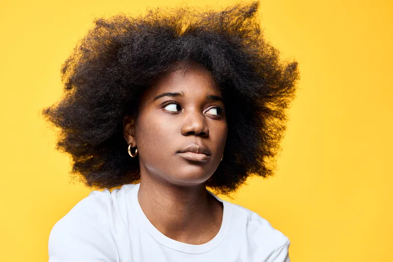 A contemplative young African American woman with voluminous natural hair against a bright yellow background, conveying emotions of curiosity and introspection