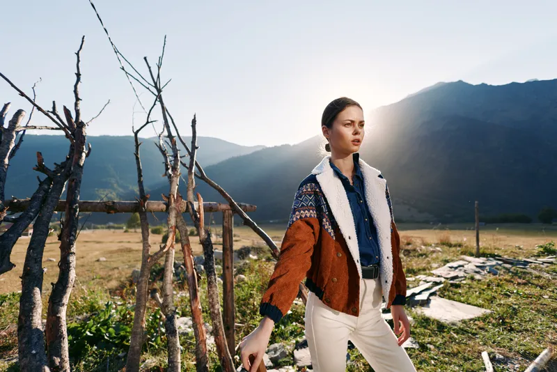 Woman in sweater and jacket standing by a rustic fence in countryside mountains at golden sunlight. Outdoor portrait, travel lifestyle with contemplative pose and natural rural scenery.