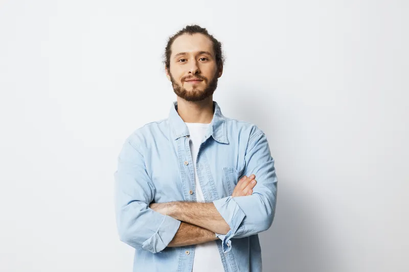 Confident young man with crossed arms in light blue denim shirt against a plain white background, exuding positivity and self assurance, ideal for corporate and personal projects