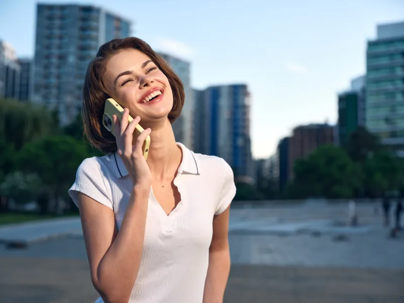A cheerful young woman is talking on a mobile phone in an urban outdoor setting. She smiles brightly, appearing confident and engaged, creating a positive business vibe.