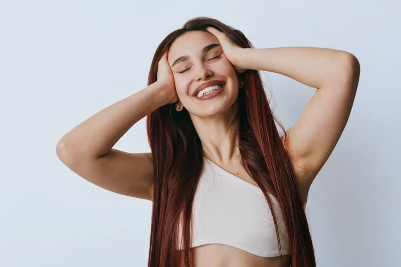 Young woman with long hair smiling with eyes closed and hands on head, wearing casual sleeveless top against plain background. Happy female portrait with relaxed joyful expression.