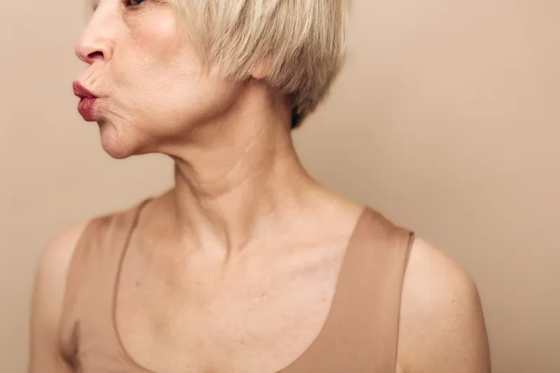 Profile of mature woman with short blonde hair wearing beige top, lips pursed as if whistling or blowing, isolated on neutral background. Concept of aging, expression, and natural beauty.