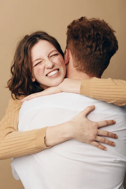 Happy young woman with curly hair hugs man wearing white shirt, smiling with closed eyes. Intimate moment of affection and love between couple in casual beige and white clothing.