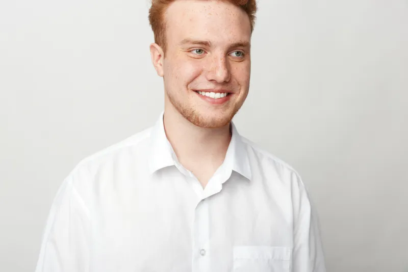 Smiling young man with red hair wearing white shirt looks away on light background. Portrait of happy male model with casual style and positive expression in studio lighting.
