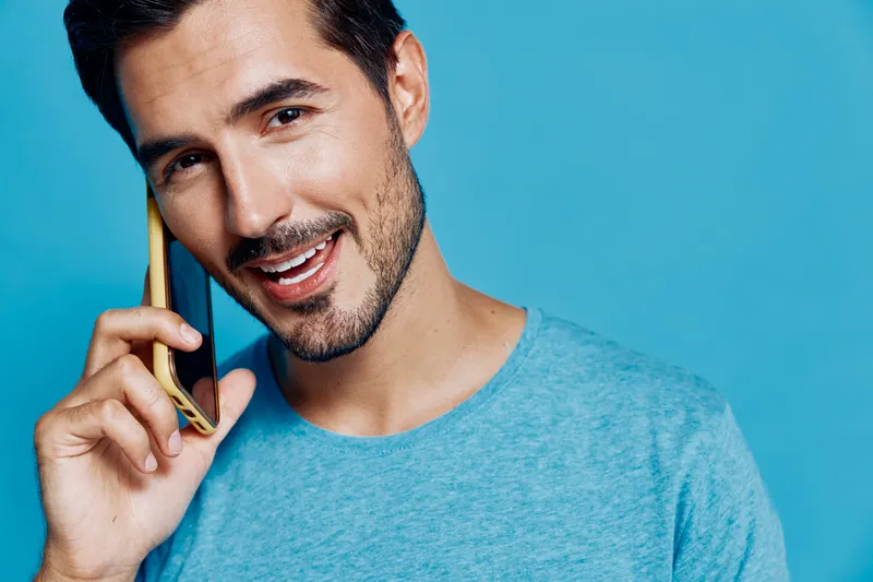 Smiling man talking on smartphone wear blue t-shirt look camera with cheerful expression isolated on bright solid blue background studio portrait. People lifestyle concept