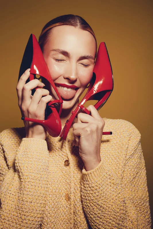 Young woman with closed eyes holds red high heel shoes near face, smiling and sticking out tongue. Fashion concept with playful mood and bright studio lighting on yellow background.