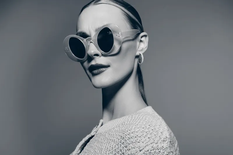 Portrait of a confident white woman with pale skin wearing large round sunglasses and a knitted top. Studio shot with neutral background, fashion and style concept.