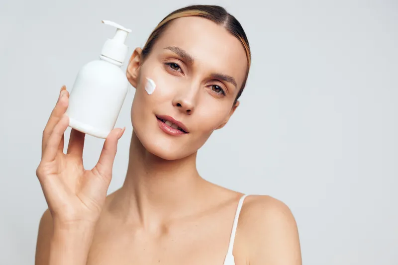 Young woman applying cream on her cheek in a soft studio setting, holding a white bottle and posing for a skincare moment with a calm, confident mood.