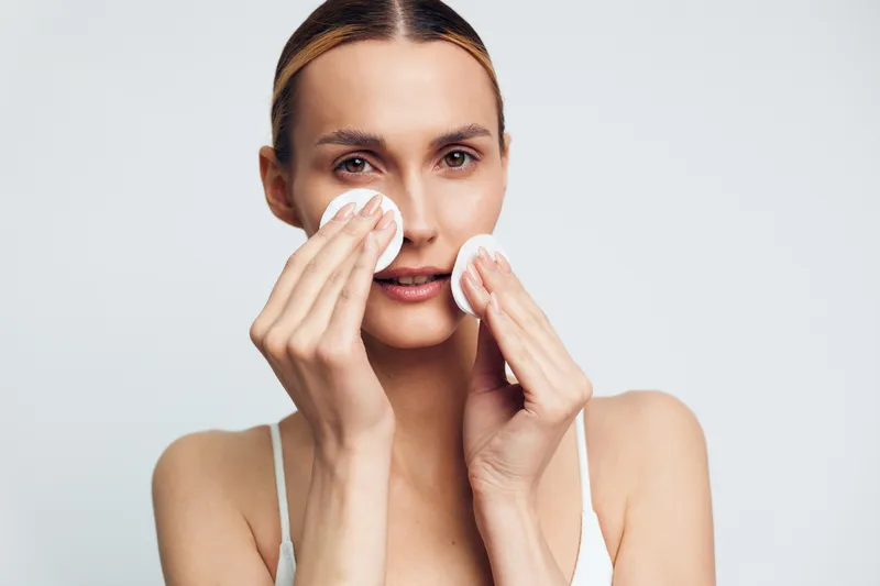A woman cleanses her face with cotton pads in a bright, minimal setting, presenting a simple skincare routine. The image conveys self care, hygiene, and clear, healthy skin.