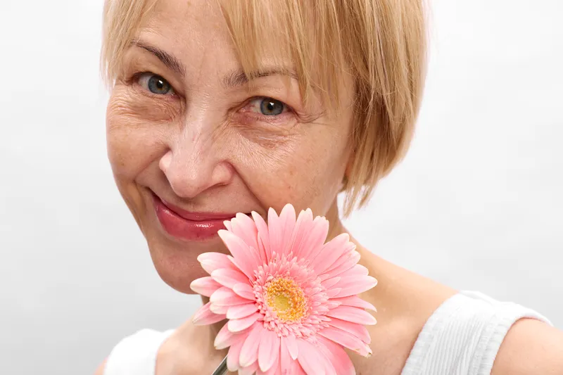 Woman portrait smiling closeup pink gerbera flower indoors, white background, springtime mood, mature blond hair, gentle face, soft light, relaxed pose