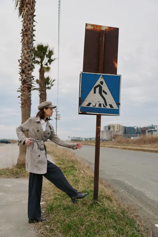 Woman in trench coat and hat stands near a rusty pedestrian crossing sign by a quiet road with palm trees. Urban outdoor scene with vintage and casual fashion concept.