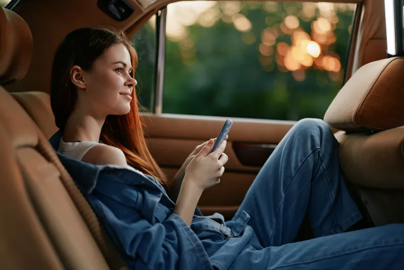 Young woman in casual clothing using smartphone in car, enjoying a moment of relaxation with a serene expression amidst a soft sunset glow.