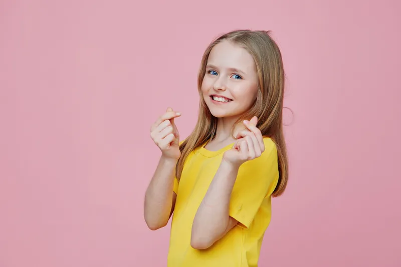 Smiling girl with long hair in a yellow shirt making heart shapes on a pink background, expressing joy and positivity, ideal for family and lifestyle themes