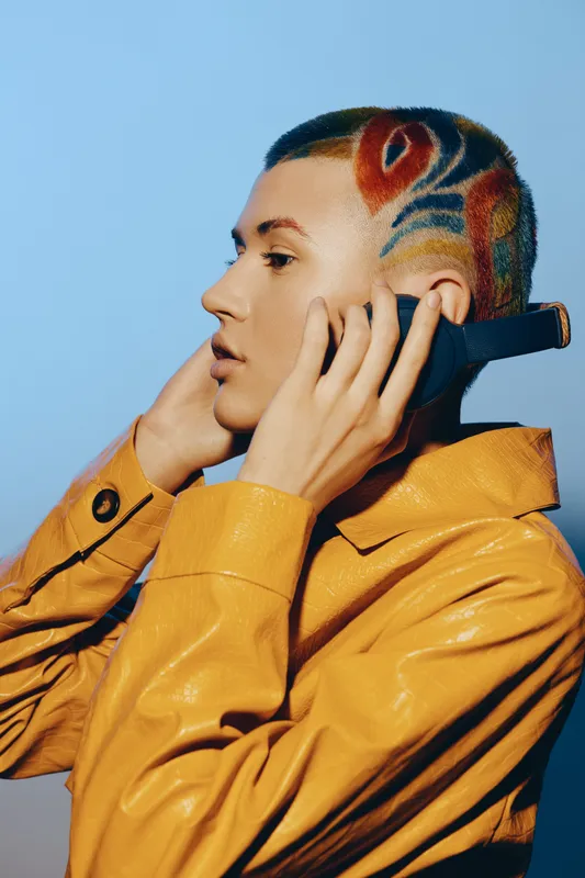 woman with colorful artistic haircut wearing yellow leather jacket listening to music on headphones, blue background, creative modern style portrait