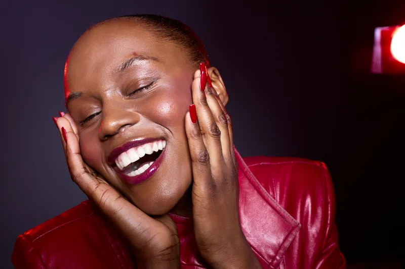 Happy African woman with dark skin and closed eyes smiling joyfully, wearing red leather jacket against dark background. Concept of beauty, happiness, and self-confidence in studio lighting.