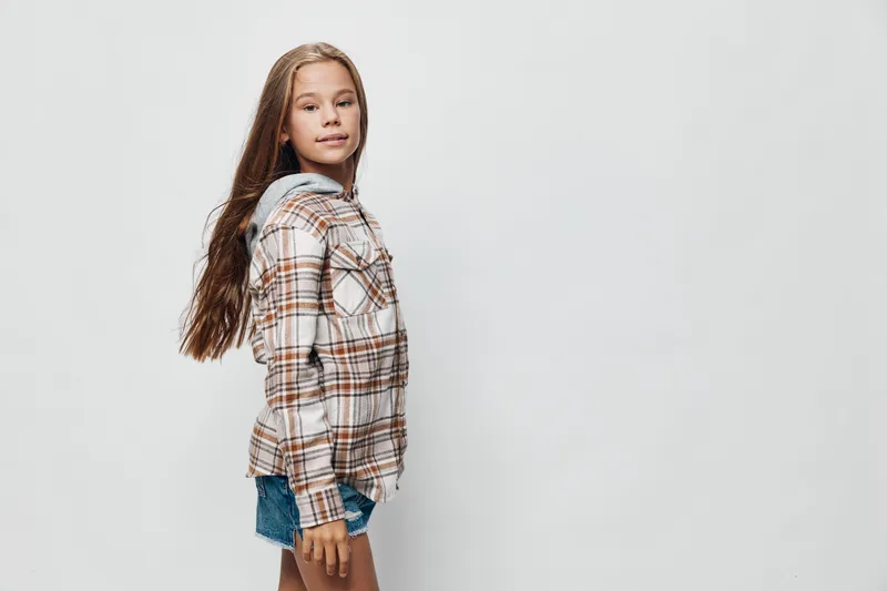 Girl with long hair wearing plaid shirt and denim shorts standing sideways on light background. Young female child in casual outfit with confident expression and natural pose.