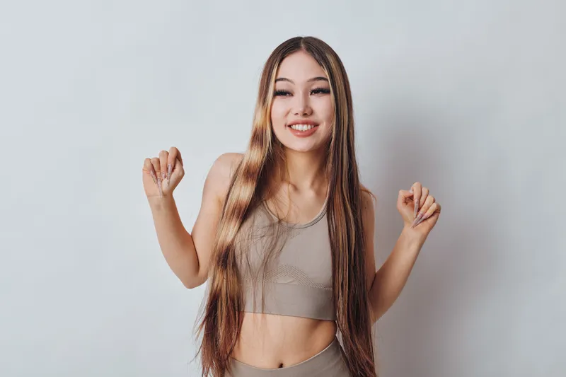 Young woman with long straight hair smiling and posing in casual sleeveless top and pants on plain light background, happy and confident female model portrait.