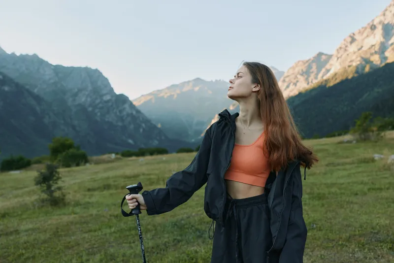 A young woman stands in a scenic valley surrounded by mountain peaks, wearing a windbreaker and hiking gear, looking up at the sky with a thoughtful gaze, capturing a moment of adventure and serenity