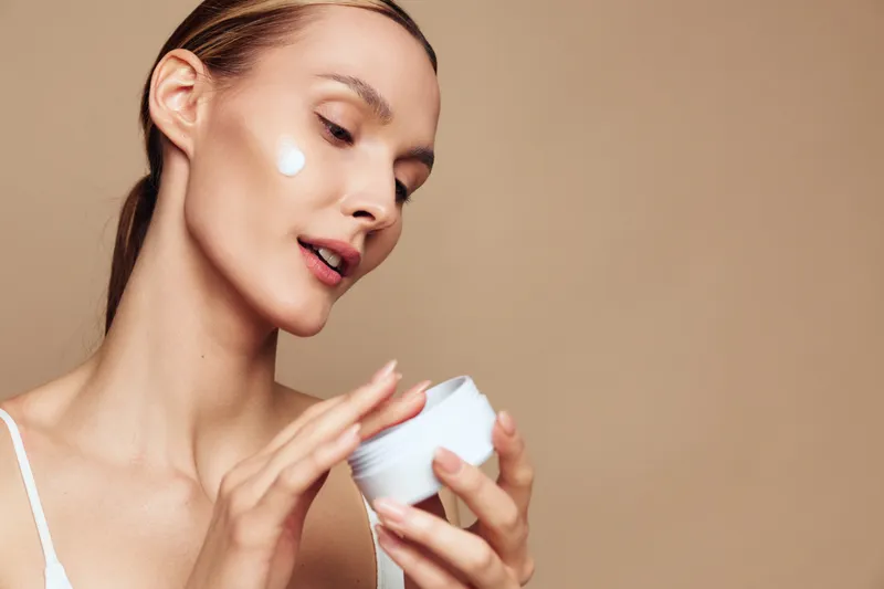 Young woman applying moisturizing cream on face, skincare routine, smooth skin, natural makeup, focused expression, neutral background, beauty concept